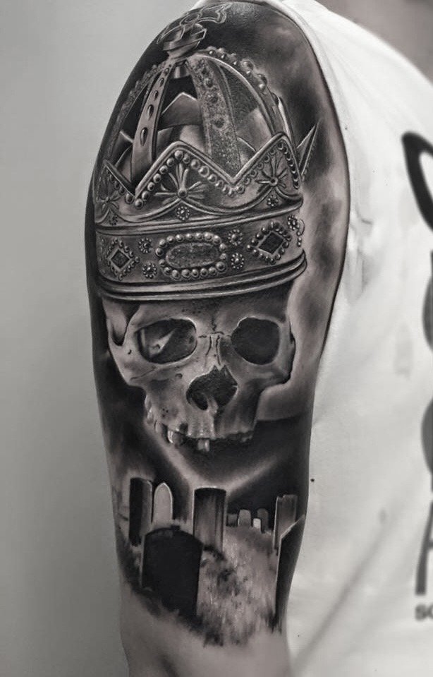 Black and grey tatovering af kranie med kongekrone og kirkegård på overarm