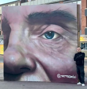 Streetart-portræt i stor skala malet med spray – realistisk øje og næse, kunstner foran værket. Fra Tattoo Mini