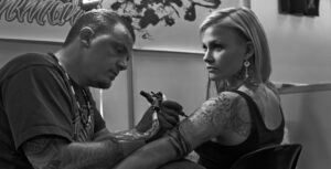 Tattoo artist intensely focused while tattooing a woman's upper arm, surrounded by detailed sketch lines and ink in a black and white setting