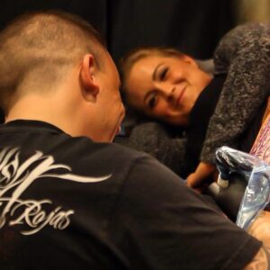 A tattoo artist working while a smiling female client looks up at him warmly, creating a moment of connection and trust