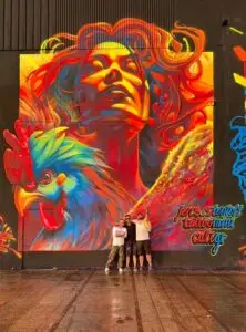 Massive mural showing a radiant woman with flowing hair in red and yellow tones, alongside a vibrant, multicolored rooster; the three artists ,Tattoo Mini - Caligr and Jens Stentoft, standing proudly in front of the artwork.