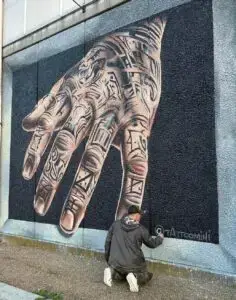 Large mural of a hyperrealistic tattooed hand extending from the wall in 3D perspective, with the artist crouched below signing the work.
