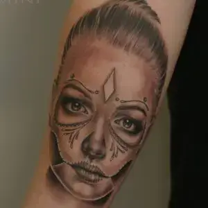 Realistic tattoo of a woman’s face styled as a sugar skull, combining portrait realism with Day of the Dead-inspired facial decorations.