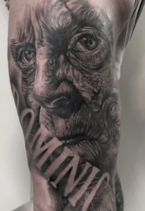 black and grey tattoo of an aged, expressive face with deeply wrinkled skin and intense eyes, with the bold name integrated into the design.
