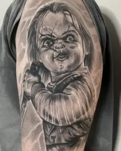 Realistic black and grey tattoo of the horror character Chucky holding a knife, inked on the upper arm with detailed facial expression, scars, and textured clothing.
