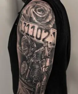 Hyperrealistic tattoo of a vintage harley davidson fork and headlight, combined with a license plate and a rose, in black and grey on an upper arm.In memory of dad