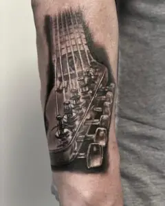 Realistic black and grey tattoo of a guitar neck and tuning pegs, tattooed along a forearm.