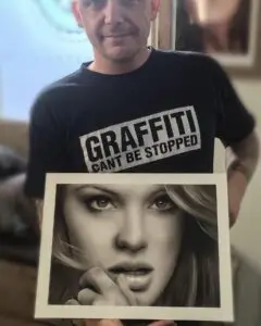 Portrait of Mini wearing a black T-shirt with the text “GRAFFITI CAN’T BE STOPPED,” holding a hyperrealistic black-and-white drawing of a woman’s face with expressive eyes and parted lips.