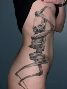 Tatovering af et dynamisk, strakt skelet i bevægelse på kvindens side og hofte, realistisk stil i sort/grå.