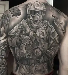 Full back tattoo of a realistic modern soldier in tactical gear holding a rifle, with helicopters above and bold lettering reading “Suck it up – Little Soldier.”