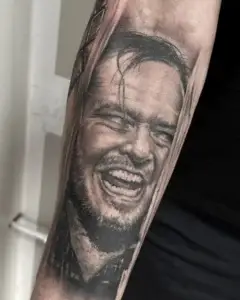 Hyperrealistic black and grey tattoo of a johnny from “the shining- with Jack Nicholson” intense laughing face, resembling a classic horror movie character, inked on a forearm with extreme detail and shading.