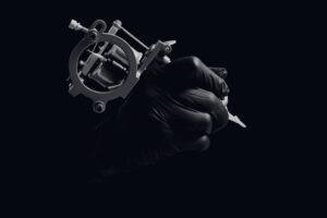 Black and white close-up photo of a tattoo machine held in a gloved hand, against a dark background with dramatic lighting.