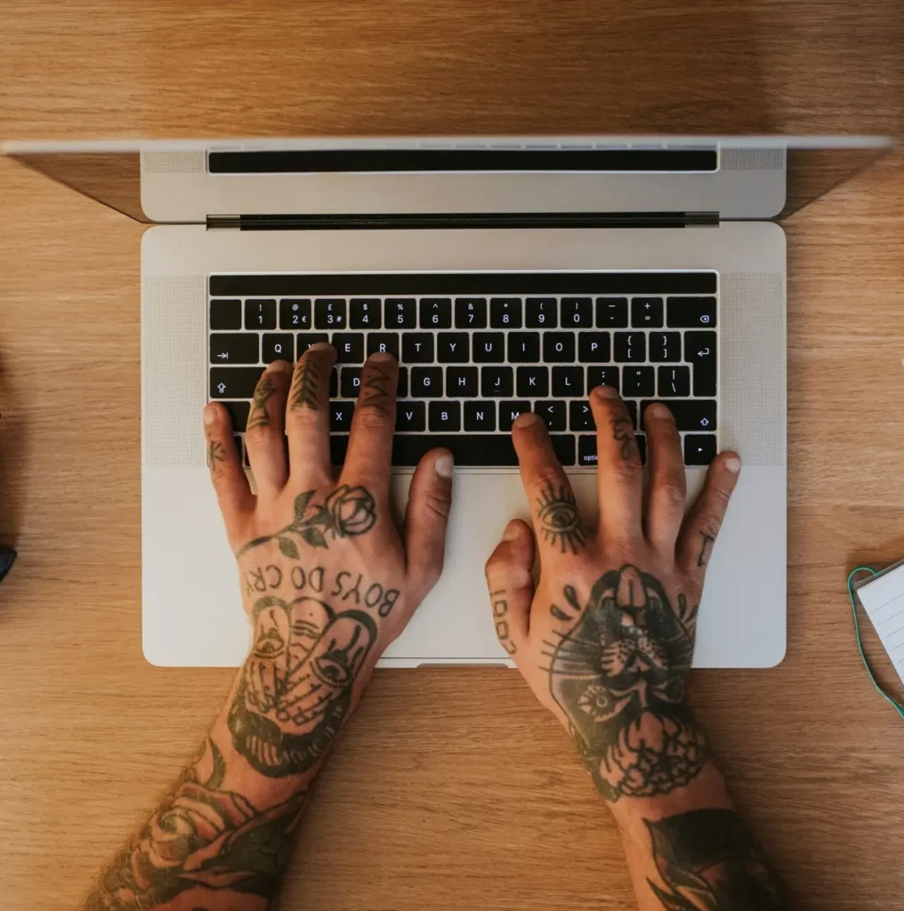 Tattoo mini hands typing on a laptop keyboard at a wooden desk with a camera, coffee cup, and notebook nearby