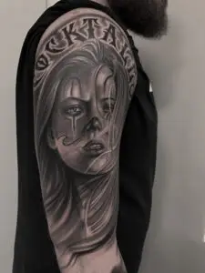 Realistisk black and grey tatovering af kvindeportræt med klovnemarkeringer og tekst på overarmen fra Tattoo Mini
