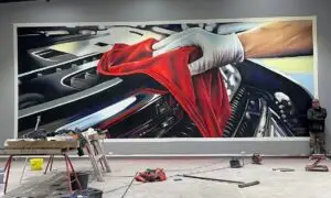 Large-scale streetart wall mural of a gloved hand polishing a car engine with a bright red cloth, painted in hyperrealistic detail inside a workshop space.with spraycans.