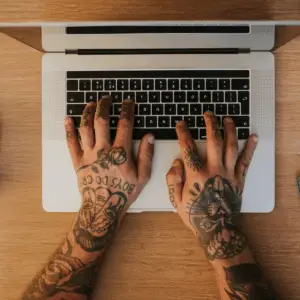 Blog om tatoveringer Tattoo mini s hands typing on a laptop keyboard at a wooden desk with a camera, coffee cup, and notebook nearby.