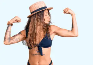 Kvinde med tatoveringer i bikini og stråhat viser styrke ved at spænde armen hos Tattoo Mini .