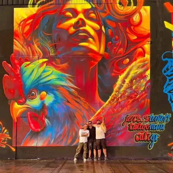 Massive mural showing a radiant woman with flowing hair in red and yellow tones, alongside a vibrant, multicolored rooster; the three artists ,Tattoo Mini - Caligr and Jens Stentoft, standing proudly in front of the artwork.